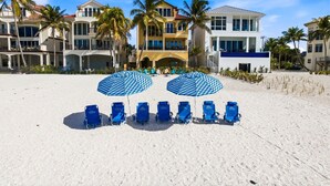 Een strand met blauwe parasols en stoelen, een groot gebouw met meerdere balkons, en palmbomen.