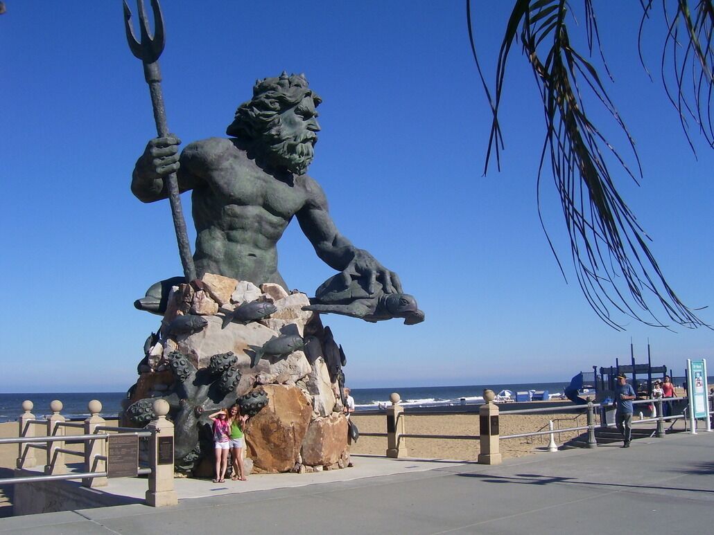 A statue of a muscular figure holding a trident, set against a clear blue sky.