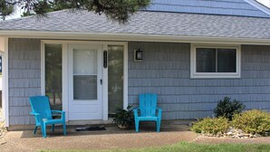 A house with a grey siding exterior, a front door, two blue Adirondack chairs, a small potted plant, and a window. - Highly rated, clean, ground floor condo w/pool; steps to beach. (Virginia Beach)