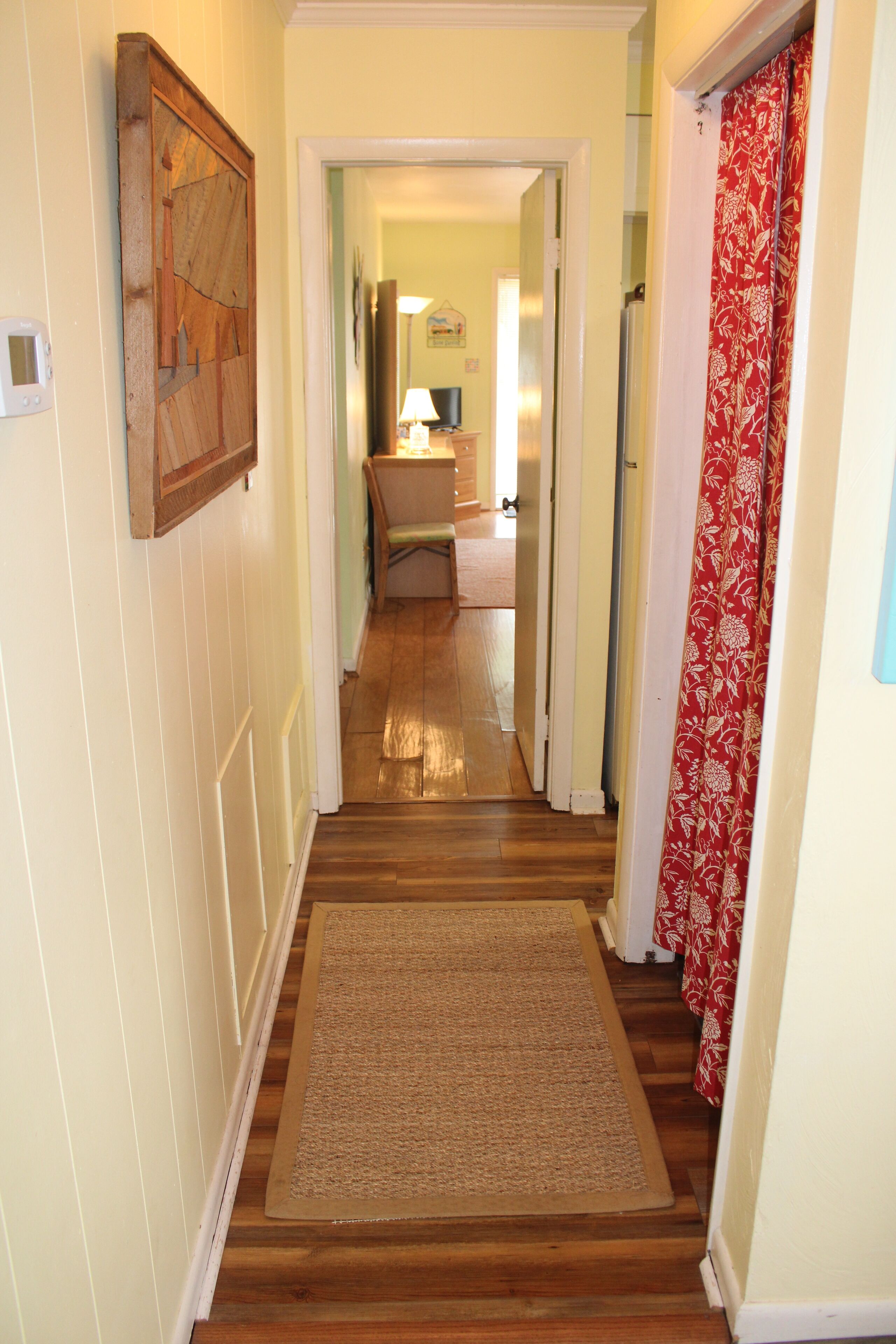 A hallway with wooden flooring, a doormat, a door with a red floral curtain, and a painting on the wall.