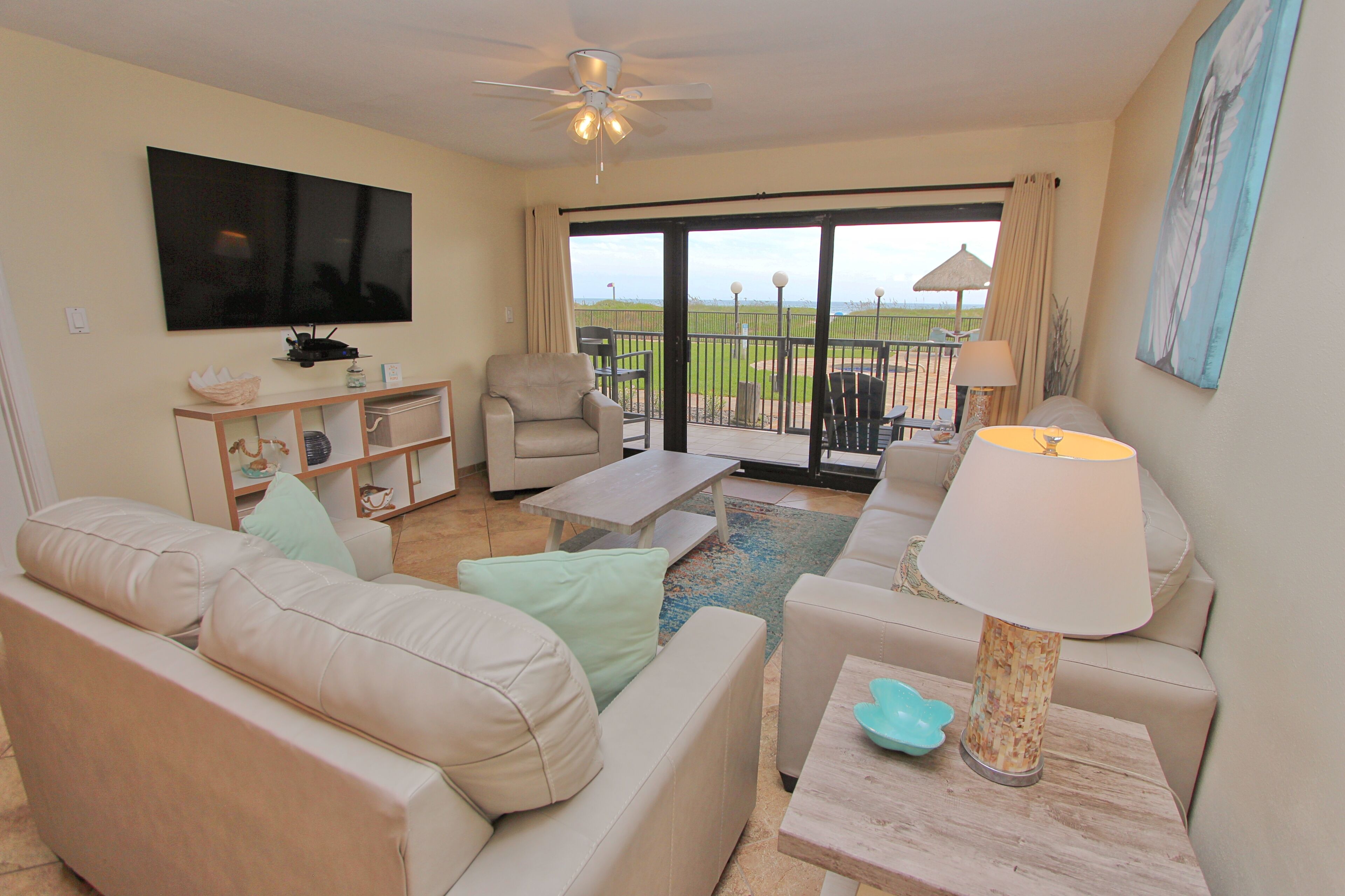 A living room with a sofa, armchair, coffee table, and a TV mounted on the wall. There is a balcony with a view of the beach.