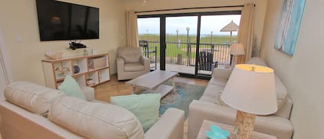 A living room with a sofa, armchair, coffee table, and a TV mounted on the wall. There is a balcony with a view of the beach.