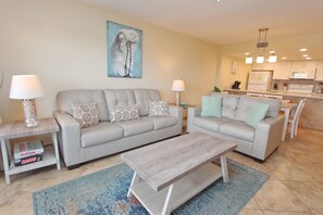 A modern living room with a grey sofa, two side tables with lamps, a coffee table, and a kitchen in the background. - Ocean's Edge - Condo 103: Spacious 2BR ground floor condo (South Padre Island)
