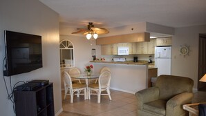 A living room with a television, a dining table with chairs, a ceiling fan, and a kitchen area with cabinets and appliances.
