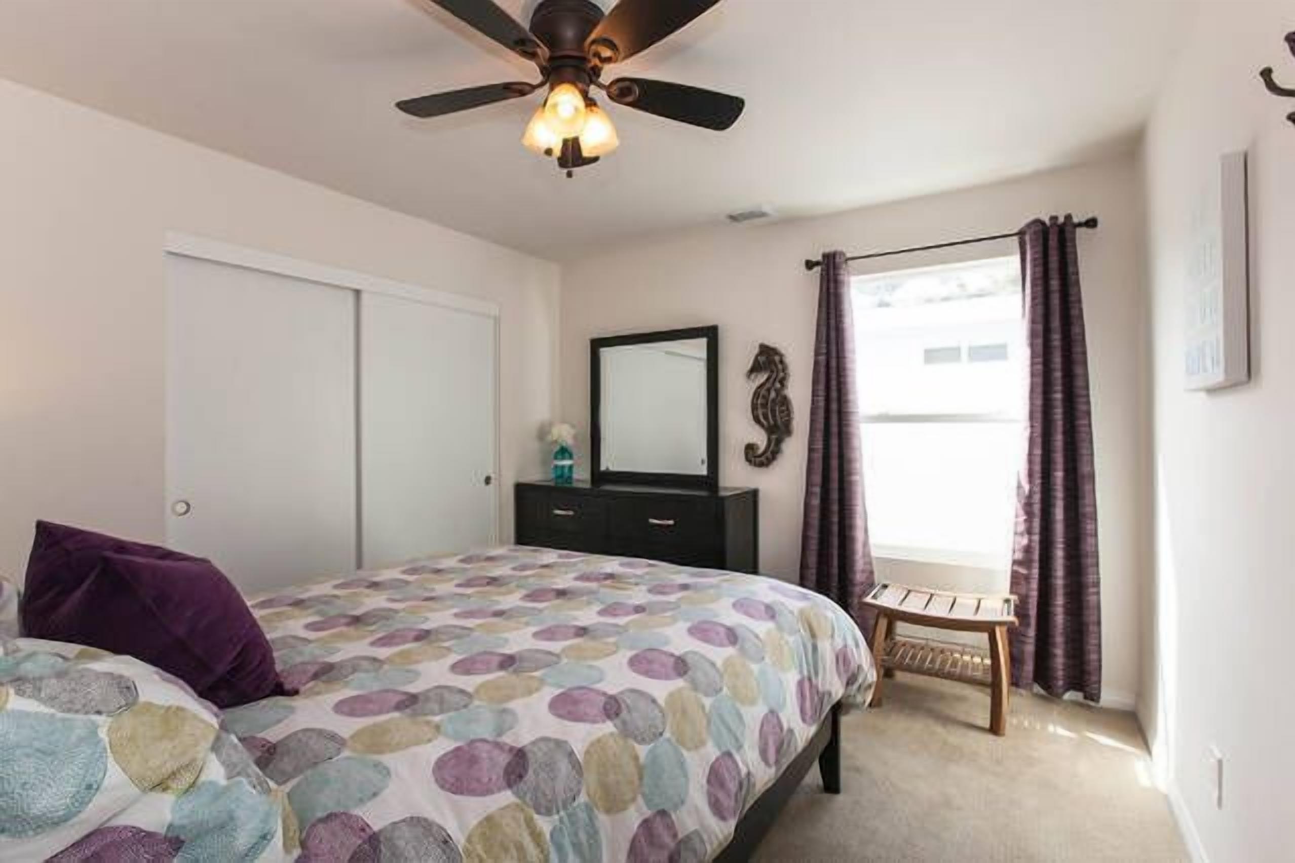 Un dormitorio con una cama, ventilador de techo, cómoda, espejo y ventana con cortinas.