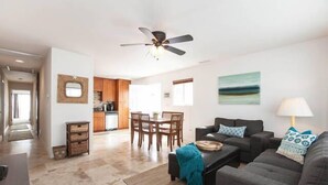 A spacious living room with a dark grey sofa, a coffee table, and a ceiling fan. There is a dining area with wooden chairs and a table, and a kitchen in the background. - Family Friendly Beach House - Rare Central Air Conditioning, Close to Town/Beach (Carlsbad)