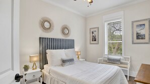 A bedroom with a large bed, two bedside tables with lamps, a window with blinds, and two wall-mounted mirrors.