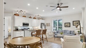 A spacious kitchen with white cabinets, a dining area with wooden chairs, a ceiling fan, and a living room with a sofa and coffee table.
