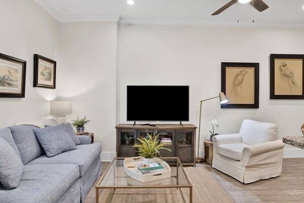 Un salón con un sofá gris, un sillón blanco, una mesa de centro de cristal y un televisor de pantalla plana sobre un mueble de madera.