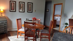 Un coin repas avec une table et des chaises en bois, une lampe, des tableaux encadrés au mur et un lit à l’arrière-plan.