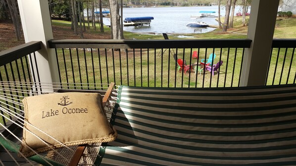 Une vue depuis un balcon, avec un hamac et un coussin portant l’inscription « lac Oconee », donnant sur une zone herbeuse où se trouvent des chaises colorées et un quai avec des bateaux.