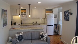 Une cuisine compacte avec des meubles blancs, un four à micro-ondes, une cuisinière et un réfrigérateur. On y trouve un canapé gris agrémenté de coussins décoratifs et une petite table avec un panier.