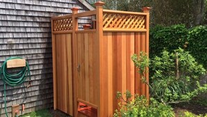 Un abri de jardin en bois, surmonté d’une treille et doté d’une petite porte, situé dans un jardin où l’on aperçoit un tuyau d’arrosage et une clôture en bois à l’arrière-plan.