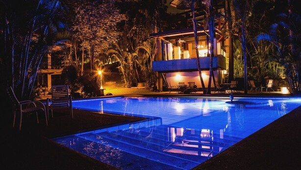 Una piscina nocturna con agua cristalina, rodeada de palmeras y un edificio con ventanas iluminadas.