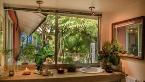 Un baño con una ventana grande, un lavamanos, un espejo y distintos artículos de aseo.