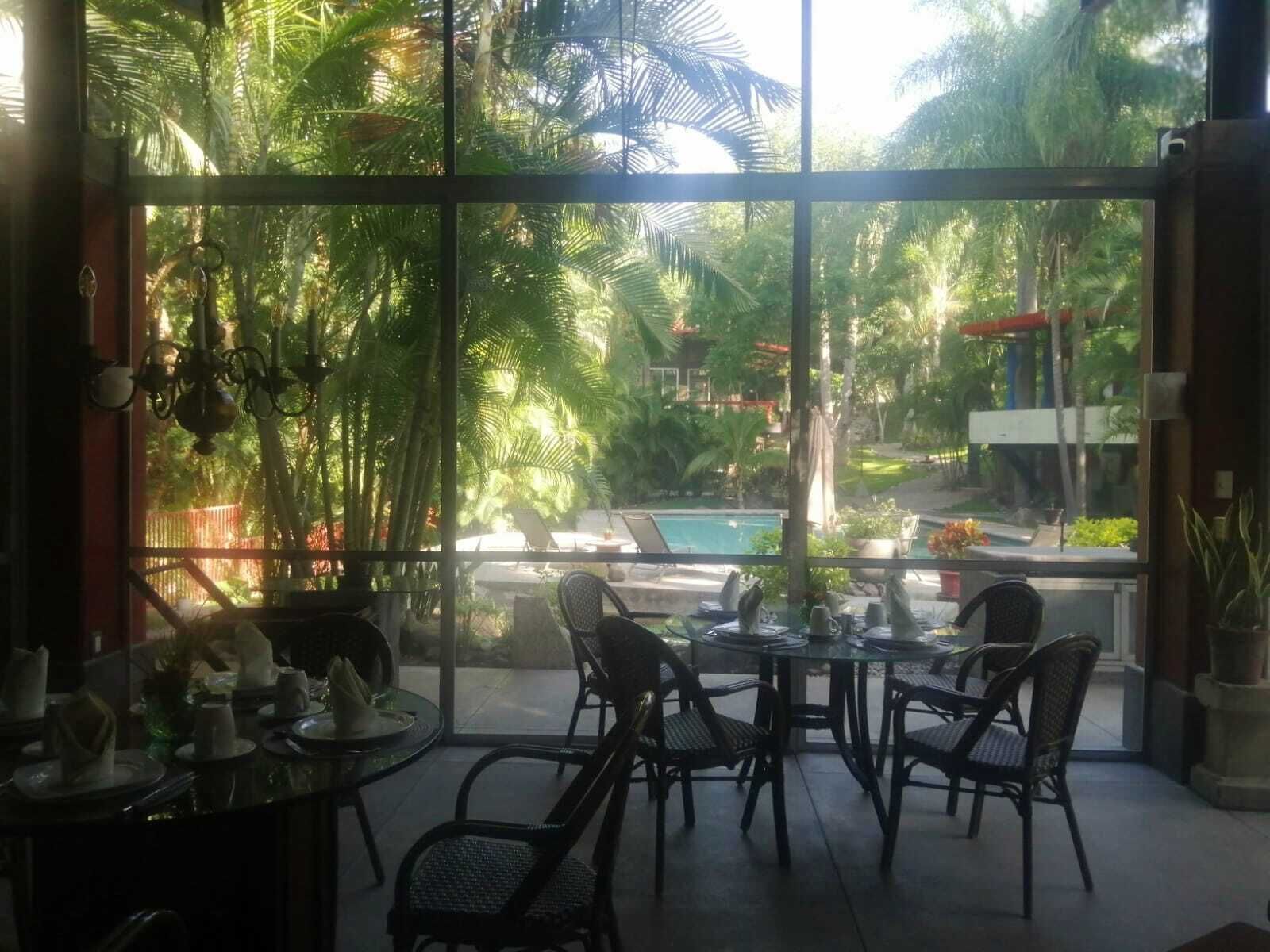A dining area with a glass table and chairs, overlooking a pool and lush greenery.