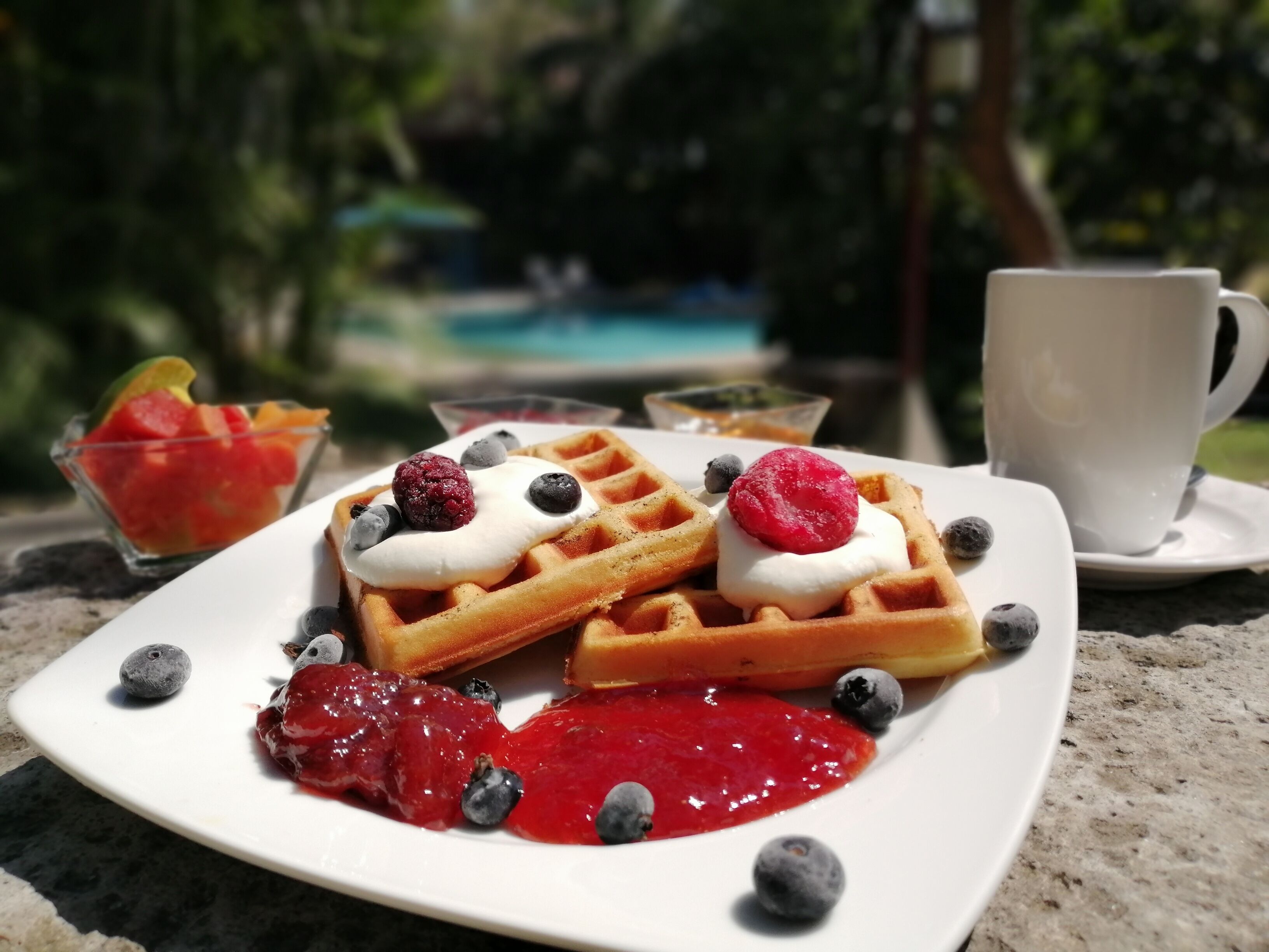 Une assiette de gaufres garnies de crème fouettée, de fruits rouges et de sirop, accompagnée d’une tasse et d’un bol de fruits.