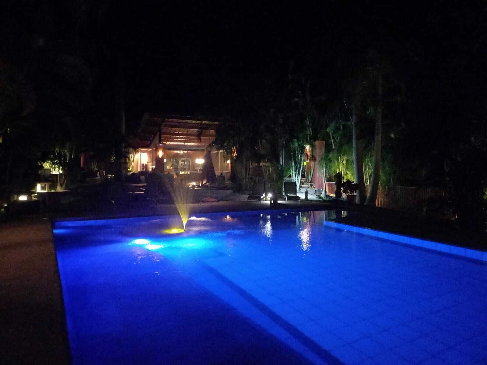 Área de piscina nocturna con una fuente, rodeada de árboles y un edificio con ventanas iluminadas.