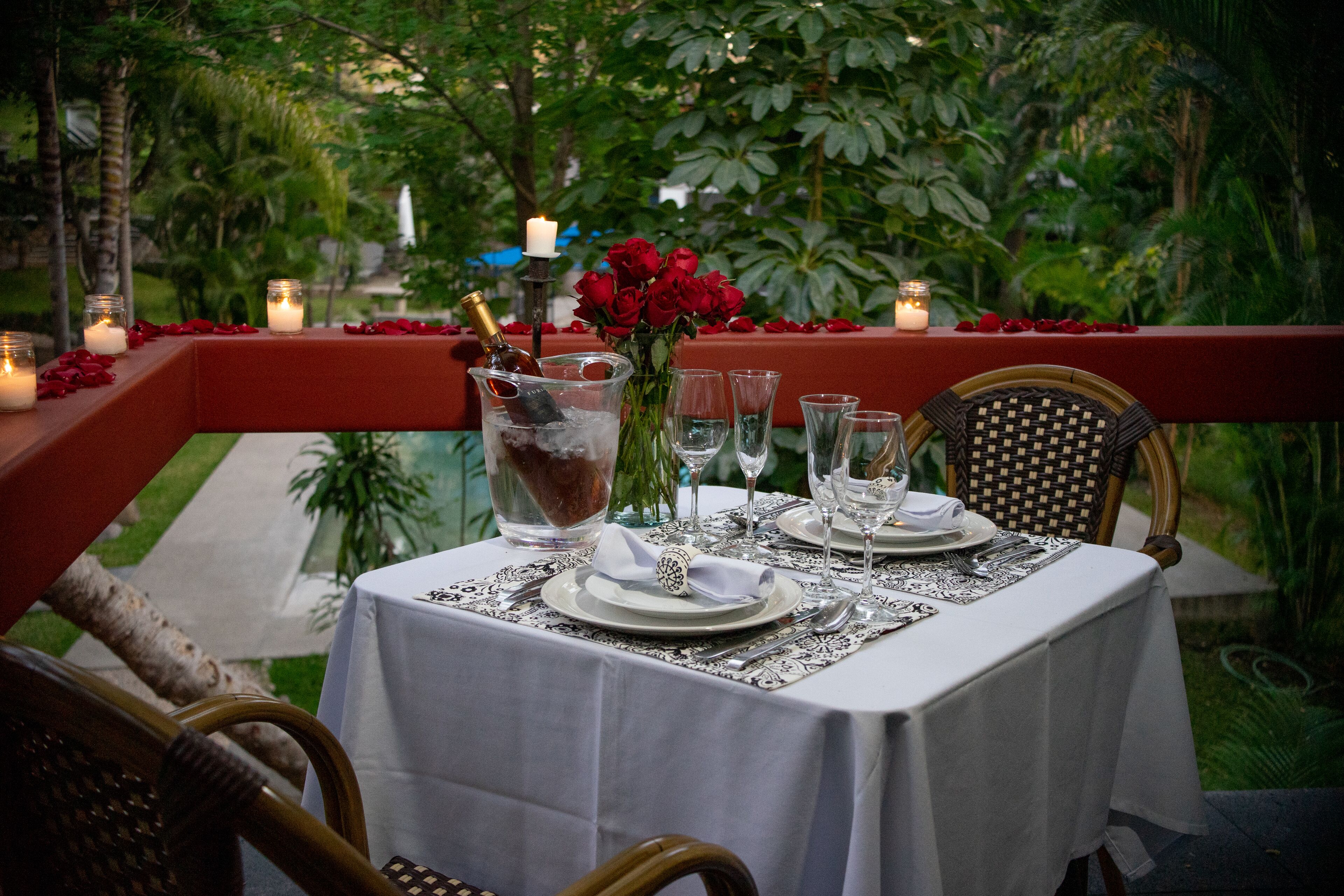 Un espace de restauration en plein air avec une table dressée pour deux personnes, entouré d’une végétation luxuriante et de pétales de fleurs rouges.
