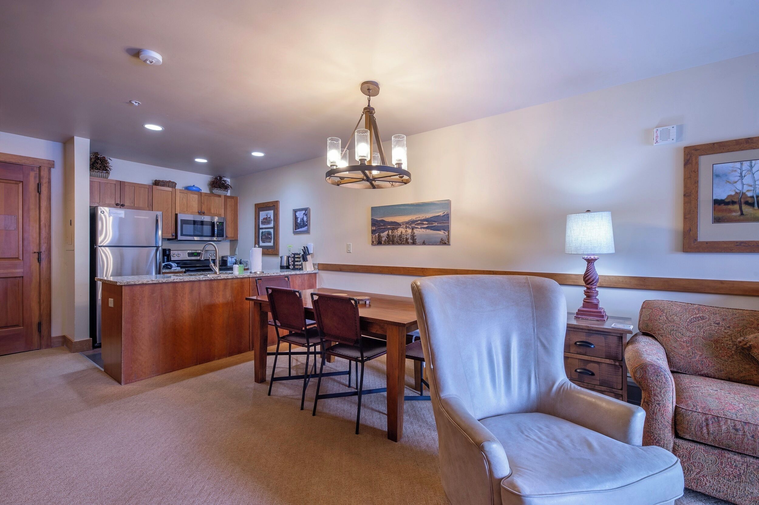 A modern kitchen with a central island, wooden cabinets, and a chandelier. There is a dining area with a table and chairs, and a living room with a sofa and a lamp.
