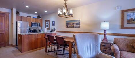 A modern kitchen with a central island, wooden cabinets, and a chandelier. There is a dining area with a table and chairs, and a living room with a sofa and a lamp.
