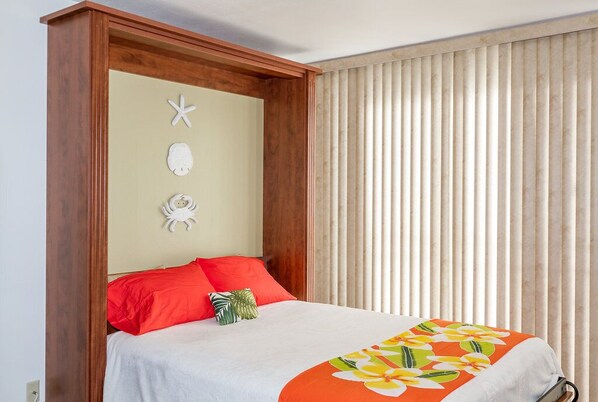 Una cama con colcha blanca y naranja estampada con flores, almohadas rojas y cabecera de madera.