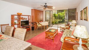 Un salon avec un téléviseur, des meubles en bois, un tapis rouge et une porte-fenêtre coulissante donnant sur un jardin.