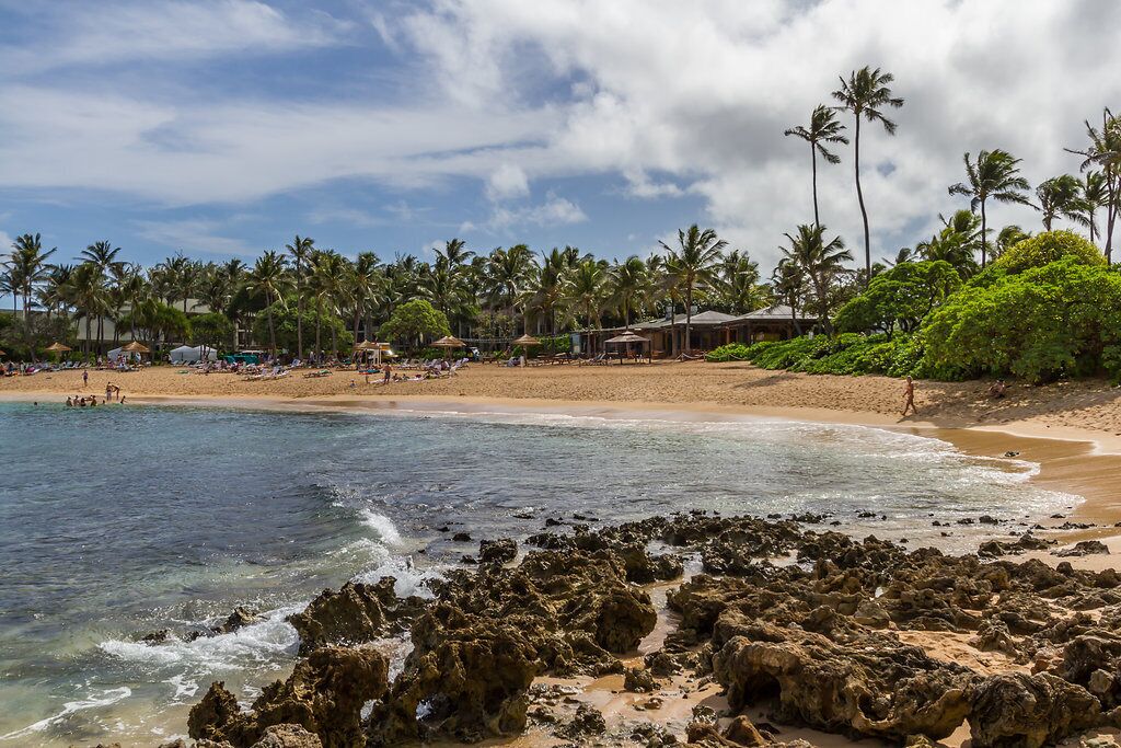 Una playa con agua cristalina, costa rocosa, palmeras y sombrillas.