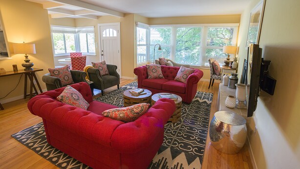 A living room with a red sofa, a green armchair, and a red armchair with patterned cushions. There is a coffee table with books and a lamp. The room has wooden flooring and a large window with blinds. - Napa Valley Retreat in Calistoga! (Calistoga)