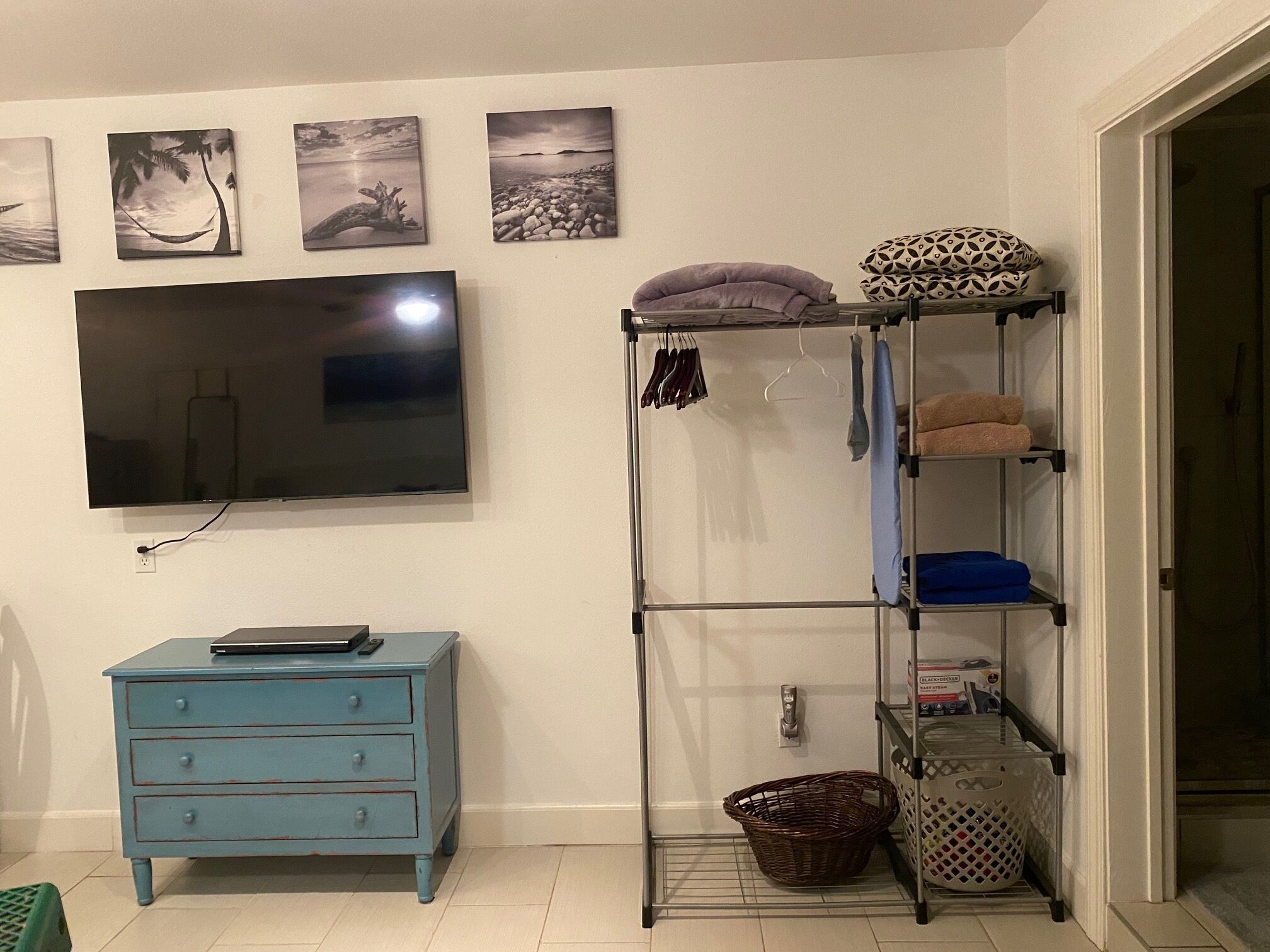 Una habitación con un televisor montado en la pared, un aparador azul, una estantería metálica con toallas y cestas, y tres cuadros colgados en la pared.