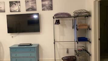 Una habitación con un televisor montado en la pared, un aparador azul, una estantería metálica con toallas y cestas, y tres cuadros colgados en la pared.