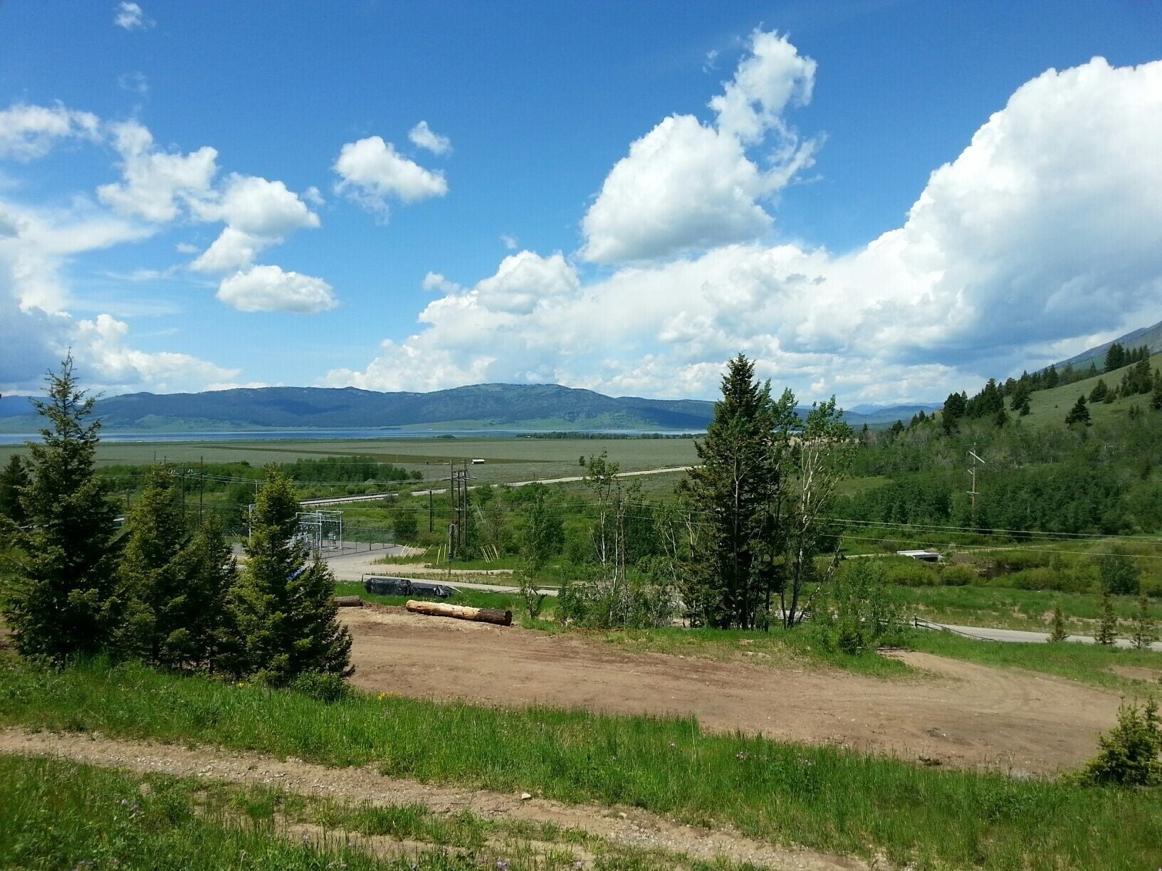 A rural landscape with a dirt road, trees, and distant mountains under a clear sky.