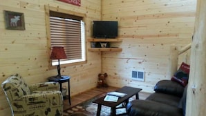 A cozy cabin interior with a wooden interior, a television mounted on a shelf, a patterned armchair, a sofa, and a small rug. - 14 miles to Yellowstone entrance. Private HOT TUB.    (Island Park)