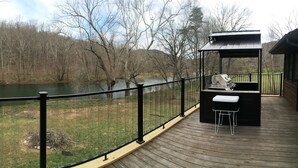 A deck with a grill and bar area overlooking a river, surrounded by a wooden deck and a metal railing. - Chalet In West Virginia on the Cacapon River. Sleeps 8 (Great Cacapon)