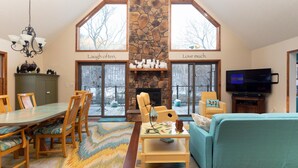 A spacious living room with a stone fireplace, a dining area with wooden furniture, and a television. - Chalet In West Virginia on the Cacapon River. Sleeps 8 (Great Cacapon)