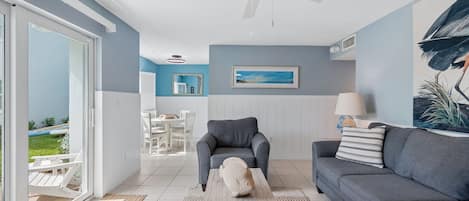 A modern living room with a grey sofa, a white coffee table, and a grey armchair. There is a ceiling fan, a framed picture on the wall, and a view of the outdoors through a sliding glass door.