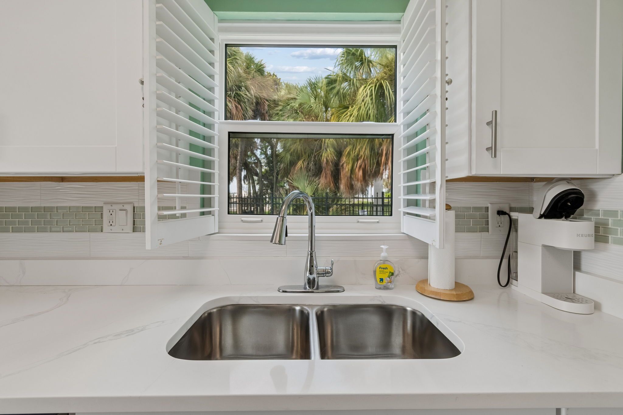 A kitchen with a double stainless steel sink, a faucet, a window with blinds, and a Keurig coffee maker.