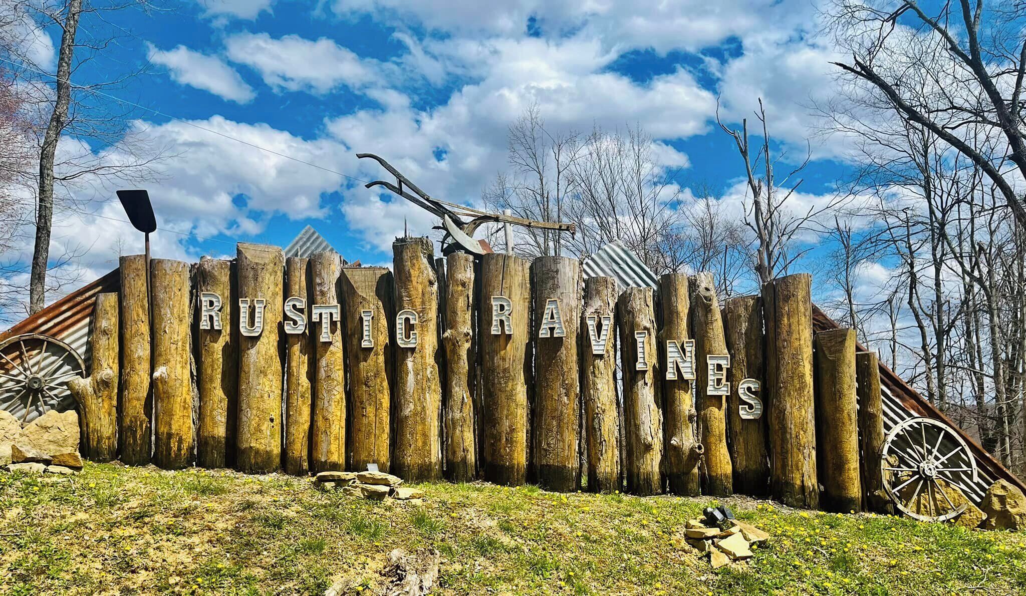 Ein rustikaler Holzzaun mit der eingravierten Aufschrift "RUSTIC RAVINES", umgeben von Bäumen und einem klaren Himmel.