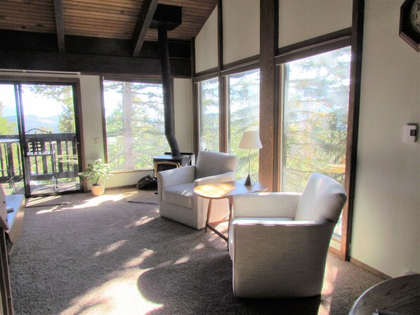 Une pièce chaleureuse avec une cheminée, deux fauteuils, une petite table et de grandes fenêtres donnant sur l’extérieur.