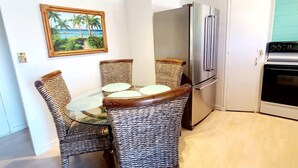 A compact kitchen with a refrigerator, oven, and a dining area with wicker chairs and a glass table. - Beautiful Lahaina Side 2 BR Condo with Beach View B7 (Honokowai)