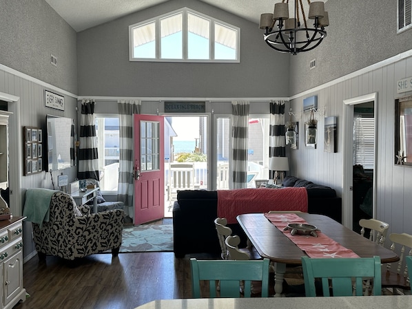 A spacious living area with a dining table, a sofa, and a view of the ocean through a large window. - Ocean View Family Home! Games, Hammocks & Chill Space Underneath. Walk to Shops! (Ocean Isle Beach)