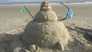 Et sandslot med gulerodsnude, knapper og pinde som arme, på en strand med oceanen i baggrunden.
