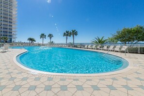Ein Pool mit Fliesen umgeben, mit Liegestühlen und Palmen, im Hintergrund ein Hochhaus.