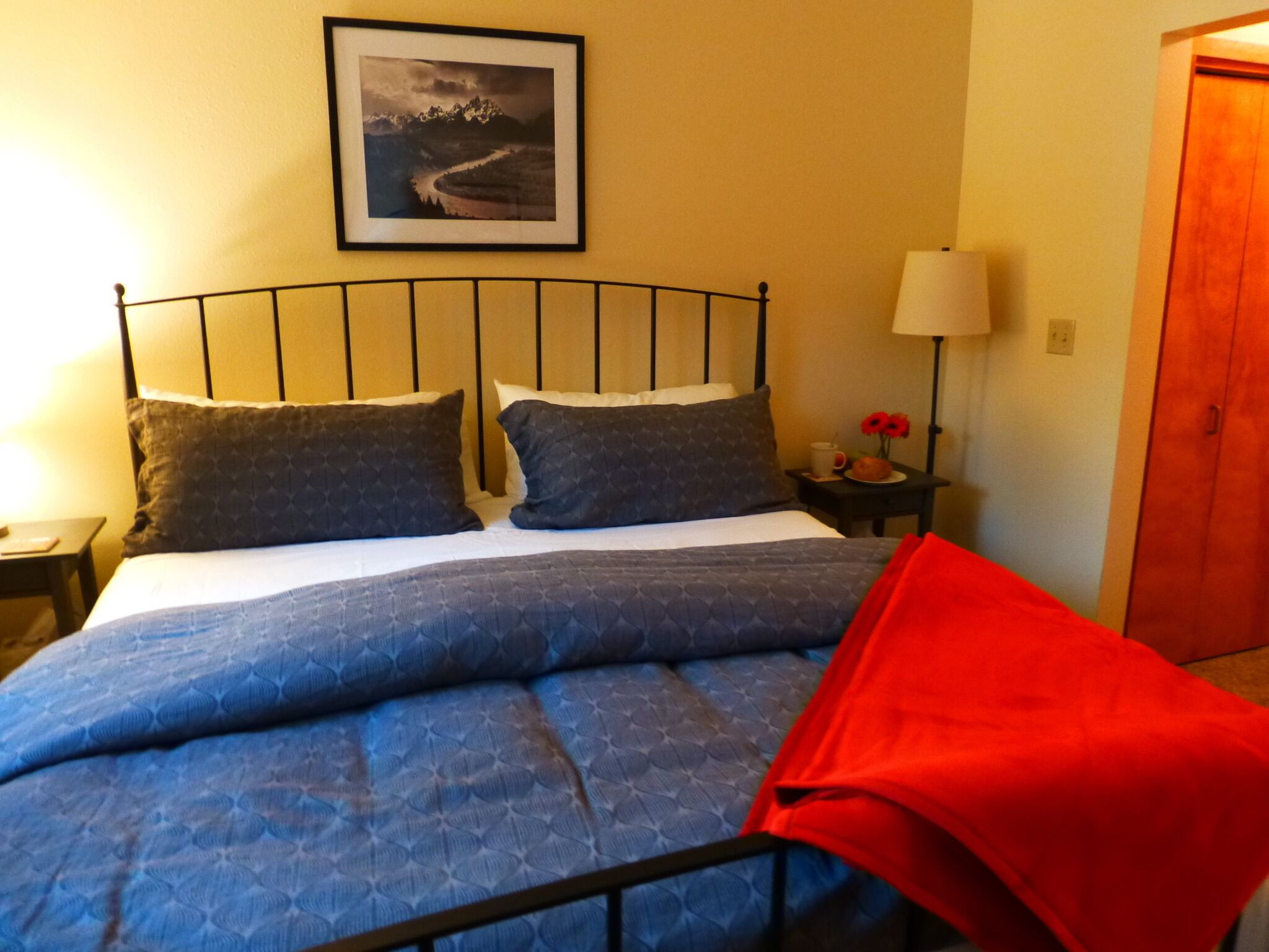 Una cama con estructura metálica, dos almohadas, una manta azul y un plaid rojo.