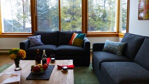 Een moderne woonkamer met een donkergrijze bank, een bijzettafeltje met een bloevas, en uitzicht op bomen door grote ramen.