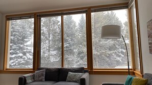Een gezellige woonkamer met een grijze bank, een klein bijzettafeltje en een lamp. Grote ramen bieden uitzicht op besneeuwde bomen.