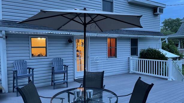 Een overdekt terras buiten met een glazen tafel, vier stoelen, een grote parasol en lichtslingers.
