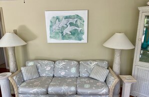 Un sofá con tapicería floral, flanqueado por dos lámparas con pantallas blancas, y una obra de arte enmarcada con delfines en la parte superior.