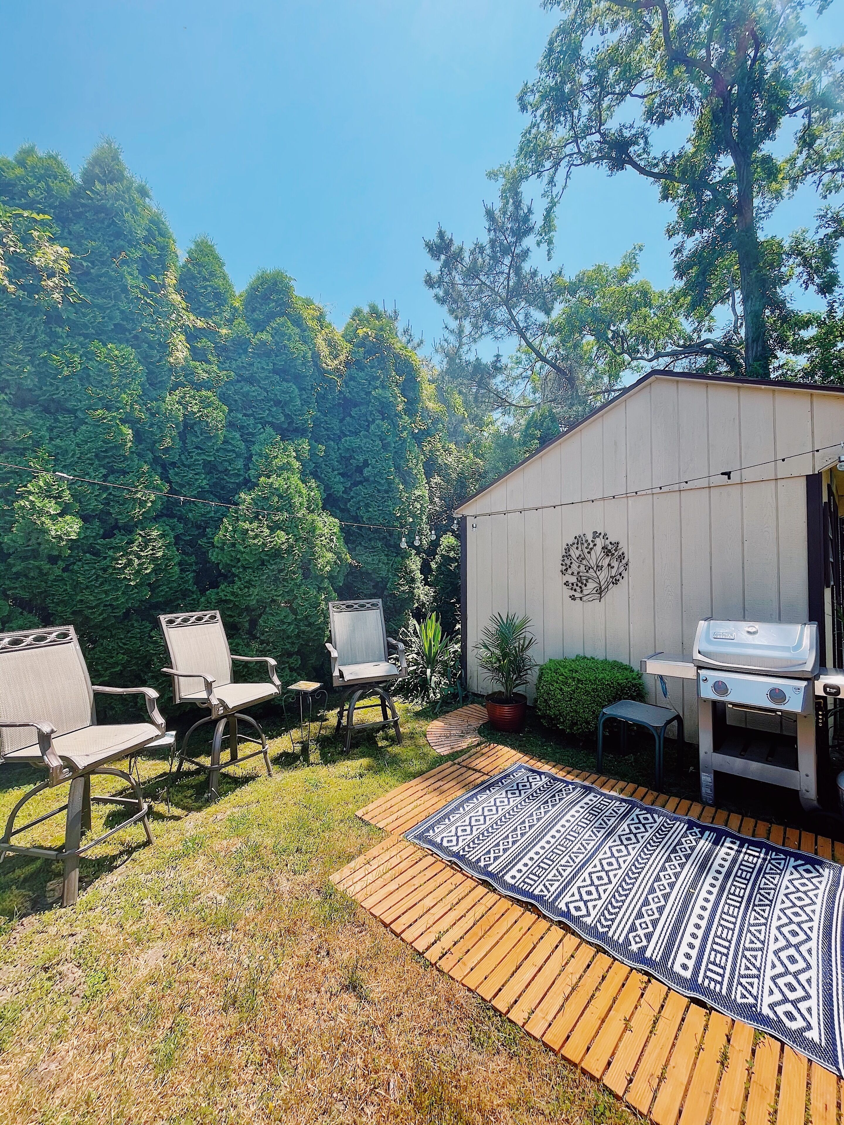 A backyard with two wicker chairs, a barbecue grill, a wooden deck, and a patterned rug.