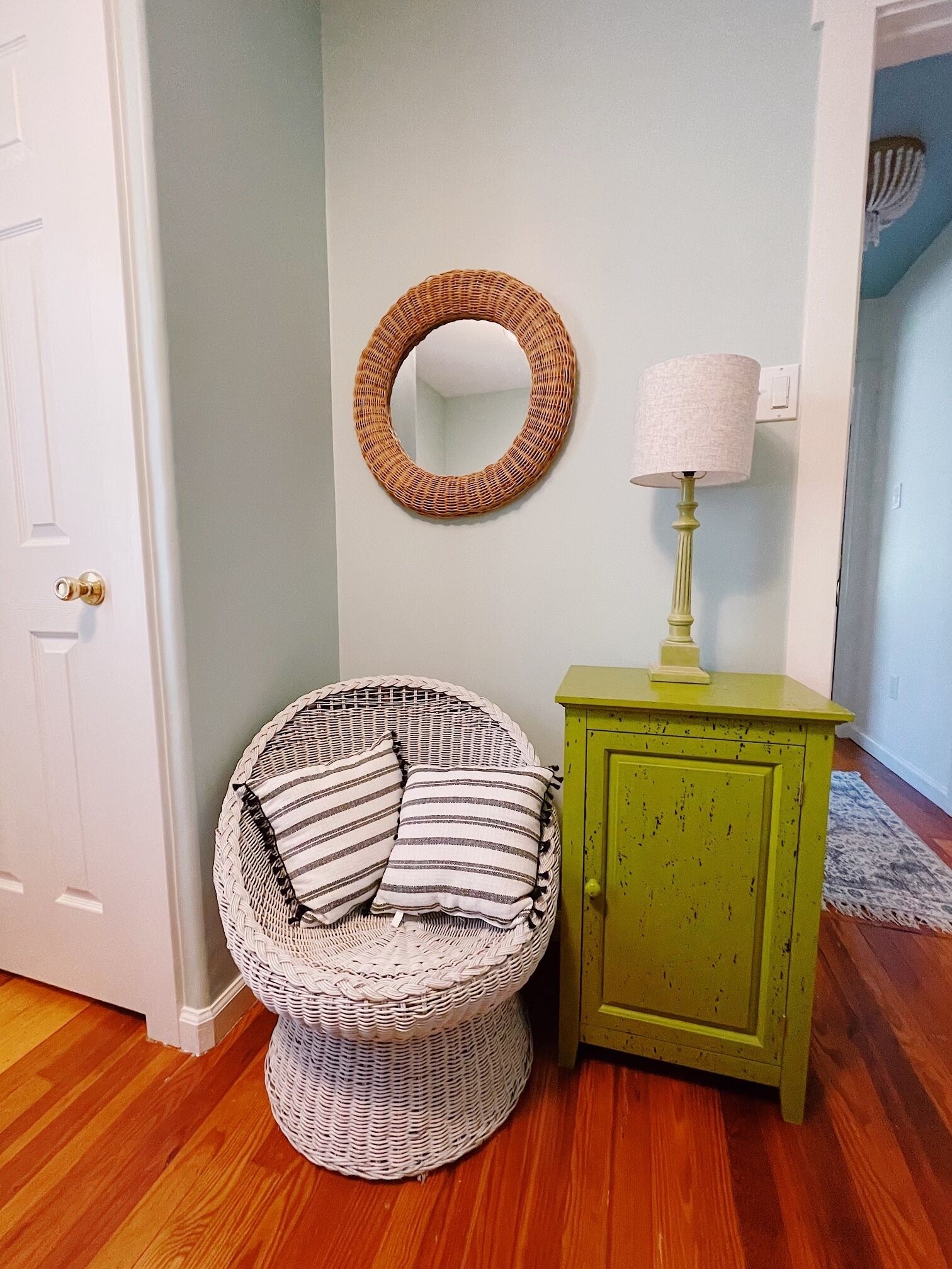 Una habitación de esquina con una silla de mimbre, un pequeño armario verde y un espejo redondo en la pared.